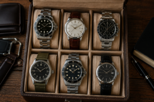 A luxury watch collection displayed in a premium leather watch case on a dark wooden desk. Six classic men's watches—including dive, dress, chronograph, and field styles—are neatly arranged in individual compartments. Surrounding accessories such as a leather wallet, key fob, notebook, and pen create a sophisticated, masculine setting that conveys timeless style, craftsmanship, and refined taste